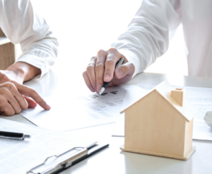 two people sitting at a table with pens and paper and a wooden house model on the table
