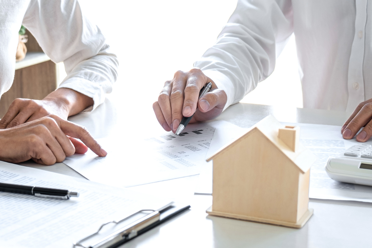 two people sitting at a table with pens and paper and a wooden house model on the table