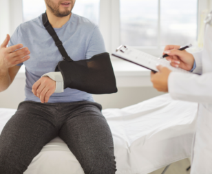 man wearing a sling sitting in a doctors office talking to a doctor with a clipboard